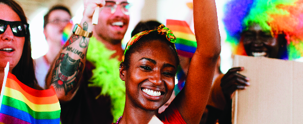 A group of diverse people wearing rainbow wigs and holding the rainbow flag.
